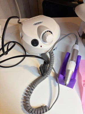 Elektrisk nagelfil - Elektrisk nagelfil med grått handstycke och spiralformad sladd. Maskinen har justerbar hastighet, fram- och bakåt-funktion samt hållare i lila plast för handstycket. Perfekt för att fila och forma naglar.