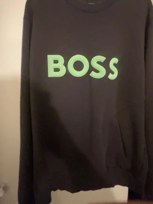 Svart sweatshirt från BOSS - Cool svart sweatshirt från BOSS med stor grön logga framtill. Tröjan har rund halsringning och långa ärmar. Perfekt för en avslappnad och trendig stil.