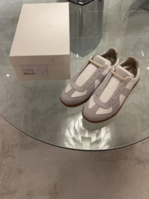 Maison Margiela Replica sneakers vita/grå - Maison Margiela Replica sneakers i vitt och ljusgrått med klassisk gummisula. Ovandelen är i en mix av mocka och läder, med snygga detaljer och minimalistisk design. Perfekta för dig som gillar stilrena och trendiga sneakers.