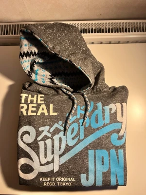 Grå Superdry hoodie med tryck - Snygg grå hoodie från Superdry med stort färgglatt tryck på bröstet i blått, vitt och gult. Huvan har ett mönstrat foder i svart, vitt och blått samt matchande dragsnören. Tillverkad i mjuk bomullsblandning för extra komfort.