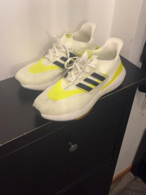 Adidas Ultraboost  - Säljer ett par snygga Adidas sneakers i vitt med gula detaljer och svarta ränder på sidan. Skorna har snörning och en chunky sula med inslag av orange undertill. Perfekta för dig som gillar sportig stil och vill sticka ut lite extra.