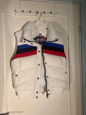 Vit dunväst med färgblock från Napapijri - Säljer en riktigt snygg vit dunväst med breda färgblock i blått, rött och svart över bröstet. Västen har hög krage, tryckknappar framtill samt två sidofickor. Perfekt för dig som vill sticka ut med sportig stil. (köpt för 800) 