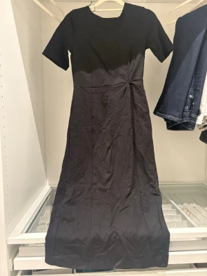 Svart långklänning med kort ärm - En stilren svart långklänning med korta ärmar och rund halsringning. Klänningen har en enkel, elegant design med lätt markerad midja och slät yta. Perfekt för dig som gillar minimalistisk stil.