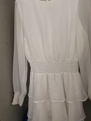 Vit volangklänning från Gina Tricot - Säljer en vit klänning från Gina Tricot med långa ärmar, markerad midja med smock och volangdetaljer nedtill. Klänningen har rund halsringning och är tillverkad i ett lätt, flowy material som ger en romantisk känsla.