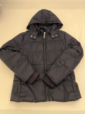 Svart dunjacka med huva från Gant - Snygg svart dunjacka från Gant med huva och dragkedja framtill. Jackan har quiltad design, ribbade muddar och tryckknappar vid axlarna. Insidan är blå med tryckt Gant-text. Perfekt för kalla vinterdagar.