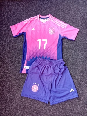 Wirtz Tysklands fotbollsset rosa/lila Adidas - Säljer ett Wirtz Tyskland fotbollsset från Adidas med matchtröja och shorts. Tröjan är rosa och lila med geometriskt mönster, nummer 17 och namnet Wirtz på ryggen. Båda delarna har broderad landslagsemblem och vita detaljer. Tillverkad i lätt polyester, perfekt för fotboll.