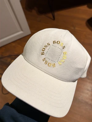 Vit keps från BOSS med gulddetaljer - Snygg vit keps från BOSS med guldfärgad logga tryckt i cirkel på framsidan. Justerbar rem baktill med metallspänne och BOSS-märke. Klassisk böjd skärm och stilren design i bomullsmaterial. Perfekt accessoar för en clean och modern look.