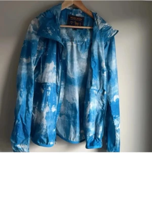 Blåvit vindjacka från Woolrich - Snygg blåvit vindjacka från Woolrich med coolt molnliknande mönster. Jackan har huva, dragkedja och lätt material som passar perfekt för blåsiga dagar. Märkesdetaljer på insidan och avslappnad passform. 