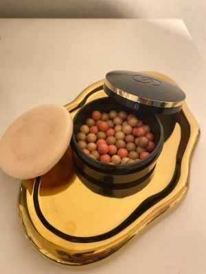 Giordani Gold bronzing pearls - Bronzer i form av pärlor i olika nyanser av brunt och persika, levereras i en rund svart burk med gulddetaljer och tillhörande mjuk appliceringssvamp. Perfekt för att ge huden en fräsch glow och solkysst look.