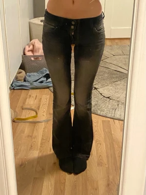Svarta bootcut jeans med knappar - Säljer ett par svarta bootcut jeans med snygg tvätt och hög midja. Jeansen har flera knappar i gylfen och utsvängda ben. Perfekta till en trendig och avslappnad stil. Materialet är stretchigt denim för skön passform.