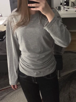 Grå långärmad topp med rynkade sidor - Snygg grå långärmad topp med rund halsringning och rynkade detaljer på sidorna. Tillverkad i mjukt bomullsmaterial som känns skönt mot huden. Perfekt att styla med jeans eller kjol för en enkel men trendig look.