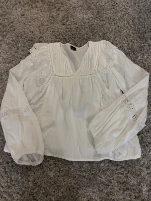Vit blus med spetsdetaljer från Gina Tricot - Söt vit blus från Gina Tricot med v-ringning och romantiska spetsdetaljer på axlar och ärmar. Blusen har långärmad design med lätt puff och resår vid ärmslut. Fint skick.