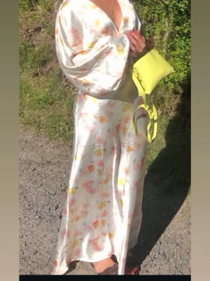 Gina Tricot klänning - Säljer en lång, glansig satinklänning med blommigt mönster i pastellfärger. Klänningen har djup v-ringning och vida ärmar som ger en avslappnad och trendig look. Perfekt för dig som gillar flowiga och eleganta plagg med färg. Ny med prislapp