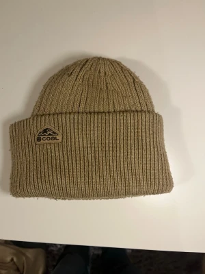 Beige ribbstickad mössa från Coal - Säljer en beige ribbstickad mössa från Coal med uppvikt kant och snyggt logomärke framtill. Klassisk modell som sitter skönt och passar perfekt när det är kallt ute. Materialet är mjukt och värmande, perfekt för dig som gillar stilrena accessoarer.
