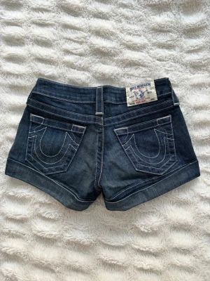 Mörkblå jeansshorts från True Religion - Säljer ett par mörkblå vintage jeansshorts från True Religion med kontrastsömmar och uppvikta benslut. Shortsen har klassiska fem fickor och en tydlig logopatch bak. Perfekta för sommaren och ger en avslappnad men snygg look. Storlek 25, säljer pga för små. Perfekt skick