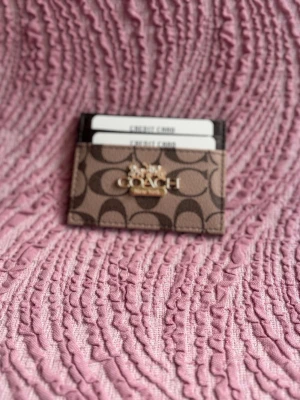 Coach brun korthållare i läder - Snygg korthållare från Coach i brunt läder med klassiskt monogrammönster och guldfärgad logga framtill. Perfekt för att organisera dina kort på ett stilrent sätt. Smidig och tunn design som enkelt får plats i fickan.