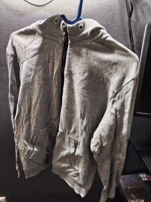 Grå hoodie från Hugo Boss - Snygg grå hoodie från Hugo Boss med dragkedja framtill och broderad logga på bröstet. Hoodien har huva, ribbade muddar och fickor på sidorna. Tillverkad i mjukt bomullsmaterial som känns skönt mot huden. Perfekt för en avslappnad och stilren look. Snöret saknas i tröjan på huvan 