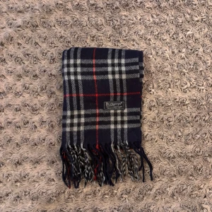 Burberry halsduk - Klassisk halsduk i mörkblått med rutigt mönster i vitt, svart och rött från Burberrys. Tillverkad i 100% lammull och har fransar i ändarna. Perfekt accessoar för att lyfta din outfit med en tidlös brittisk stil.