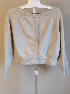 Beige croppad stickad tröja - Säljer en beige croppad stickad tröja med bred halsringning och långa ärmar. Tröjan har ribbade muddar vid ärmslut och nederkant samt en dekorativ söm framtill. Perfekt för lager på lager och enkel att styla.