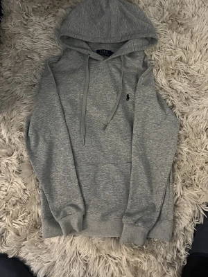 Grå hoodie från Polo Ralph Lauren - Snygg grå hoodie från Polo Ralph Lauren med klassisk huva och dragsnören. Sitter som S Tröjan har en liten broderad logga på bröstet och en stor magficka. Tillverkad i mjukt bomullsmaterial som känns skönt mot huden. Perfekt för en avslappnad och stilren look.