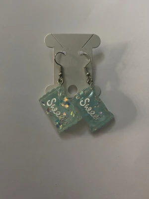 Blå örhängen med texten Sweet - Unika örhängen i ljusblå plast med skimrande glitterdetaljer och texten 'Sweet' på varje hänge. De har en lekfull, fyrkantig form som påminner om godisförpackningar och hängs på örat med silvriga krokar. Perfekta för dig som gillar söta och färgglada accessoarer.