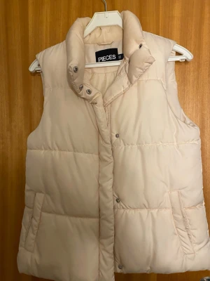 Beige dunväst från PIECES - Snygg beige dunväst från PIECES med hög krage och tryckknappar framtill. Västen har två fickor på sidorna och quiltad design som ger en puffig look. Perfekt för lager på lager och enkel att matcha med olika outfits.