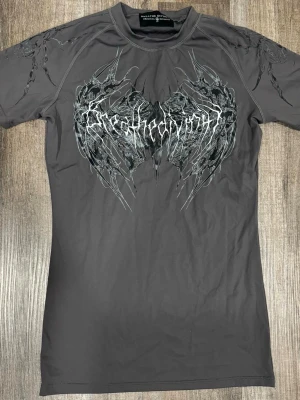 Breathedivinity L - Säljer en grå t-shirt från breathedivinity med ett stort grafiskt tryck i svart och vitt på bröstet. T-shirten har korta ärmar och en slim passform. Perfekt för dig som gillar edgy streetwear och unika prints.