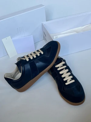 Marinblå sneakers från Maison Margiela - Snygga marinblå sneakers från Maison Margiela med beige snörning och klassisk gummisula. Ovandelen är i en mix av mocka och läder för en lyxig känsla. Diskret logga på plösen och stilren design som passar till det mesta.