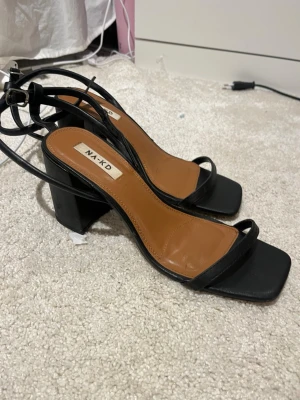 Svarta klackar från NA-KD - Stilrena sandaletter från NA-KD med svart remdesign och fyrkantig tå. Skorna har en bred blockklack och brunt innersula i syntetmaterial. Remmarna knyts runt vristen för en trendig look. Perfekta för dig som gillar minimalistisk stil och vill ha något som sticker ut.