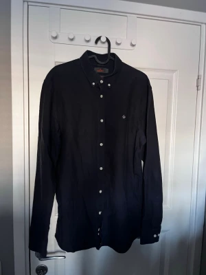 Svart skjorta från Morris - Stilren svart skjorta från Morris med klassisk krage och vita knappar. Skjortan har lång ärm och diskret logga broderad på bröstet. Perfekt för dig som gillar en clean och tidlös look.