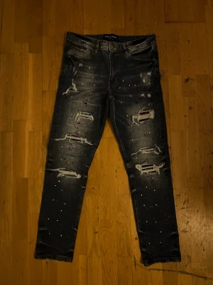 Blå distressed jeans med stänk - Säljer ett par mörk blå slim fit jeans från supply demand med slitningar och hål på framsidan samt vita färgstänk för en edgy look. Jeansen har fem fickor och klassisk knappgylf. Perfekta för dig som gillar streetwear och vill sticka ut.