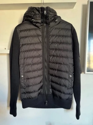 Monckler cardigan  - Snygg svart dunjacka från Moncler med quiltad front och stickade ärmar. Jackan har huva, dragkedja framtill och Moncler-logga på ärmen. Perfekt för kyliga dagar och ger en sportig men stilren look.