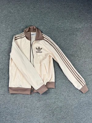 Beige Adidas Original tröja - Snygg beige track jacket från Adidas med bruna detaljer och klassiska ränder längs ärmarna. Jackan har hög krage, dragkedja framtill och två fickor med dragkedja. 
