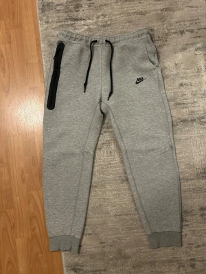 Gråa Nike mjukisbyxor med dragkedja - Snygga grå mjukisbyxor från Nike med svart logga på vänster lår, svarta dragsnören i midjan och en lång svart dragkedjeficka på höger sida. Byxorna har ribbade muddar vid bensluten och är tillverkade i mjukt material för maximal komfort.