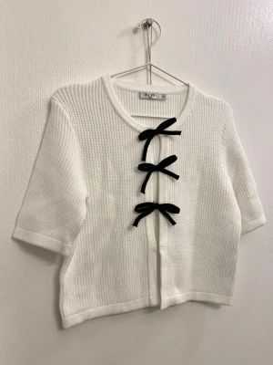 Vit kofta med svarta rosetter  - Superfin vit ribbstickad kofta från NA-KD med kort ärm och tre svarta rosetter framtill. Koftan har rund halsringning och är gjord i ett mjukt stickat material. 