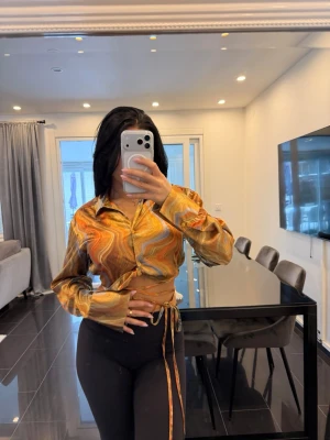 Mönstrad omlott satinskjorta i orange toner - Snygg skjorta med swirl-mönster i orange, gult och brunt. Skjortan har omlott-detalj med knytband i midjan och långa ärmar. Perfekt för dig som vill sticka ut med färg och form.