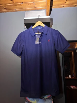 Marinblå pikétröja från Polo Ralph Lauren - Klassisk marinblå pikétröja från Polo Ralph Lauren med kort ärm och krage. Tröjan har en liten röd broderad logga på bröstet och knäppning framtill. Perfekt för en stilren och avslappnad look.