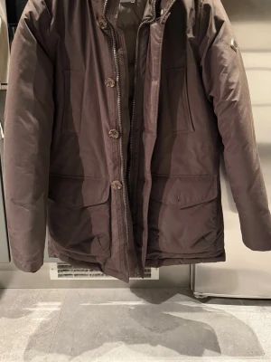 Brun parkas från J. Lindeberg - Snygg brun parkas från J. Lindeberg med dragkedja och knappar framtill. Jackan har två stora fickor med lock och en diskret logga på ärmen. Perfekt för kalla dagar och har en klassisk, rak passform. Materialet är slitstarkt och ger en stilren look.