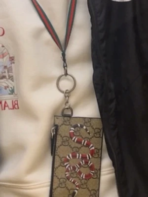 Gucci plånbok med orm och monogram - Snygg plånbok från Gucci med klassiskt GG-monogram i beige och brunt, dekorerad med en röd och vit orm på framsidan. Plånboken har en praktisk rem i grönt och rött för att hänga runt halsen. Perfekt accessoar för dig som gillar lyxiga detaljer.