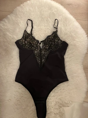 Svart spetsbody med djup urringning - Snygg svart body med djup v-ringning och tunna axelband. Överdelen är i transparent spets med blommönster och resten i slätt tyg. Perfekt för att känna sig extra fin. Tryckknappar i grenen för enkel på- och avtagning.