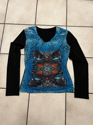 Mönstrad långärmad topp i blått och svart - Cool långärmad topp med svart ärmar och blått zebramönster på framsidan. Mittpartiet har ett färgstarkt tryck i orange, svart och blått med glittriga detaljer. Rund halsringning och figurnära passform. Perfekt för dig som gillar unika prints.