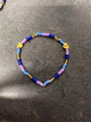 Färgglatt pärlarmband med hjärtan storlek M  - Armband med pärlor i blått, lila, rosa och guld. Två guldfärgade hjärtan ger extra detalj. 