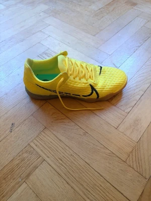 Gula Nike inomhusfotbollsskor - Använda en gång. Säljer ett par snygga fotbollsskor från Nike i klargult med svart swoosh på sidan och tå. Skorna har snörning och en unik sula med turkosa detaljer för extra grepp. Ovandelen är i syntetmaterial med textur för bättre bollkontroll.