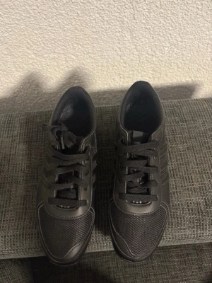 Svarta Adidas La trainers ll - Snygga svarta Adidas sneakers med klassiska tre ränder på sidan. Skorna har ovandel i mesh och syntetmaterial för bra andningsförmåga och komfort. Yttersulan har gummidobbar för extra grepp. Perfekta för en sportig och avslappnad stil.