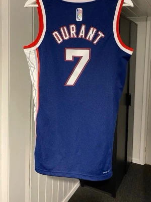  Brooklyn Nets Kevin Durant Linné  - Säljer en blå baskettröja från Nike med Brooklyn Nets tryck och nummer 7 för Kevin Durant. Tröjan har röda och vita detaljer, breda axelband och är tillverkad i ett lätt, ventilerande material. Perfekt för basket eller streetwear.