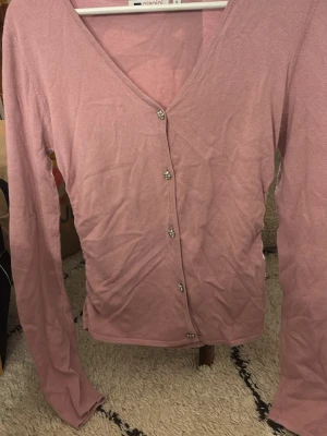 Rosa kofta med blommiga diamant knappar - Söt ljusrosa kofta från Gianini med långa ärmar och v-ringning. Koftan har dekorativa blommiga knappar med små glittrande stenar som ger en extra detalj. Tunn och mjuk i materialet, perfekt att slänga över en topp. Scrunch på sidorna.