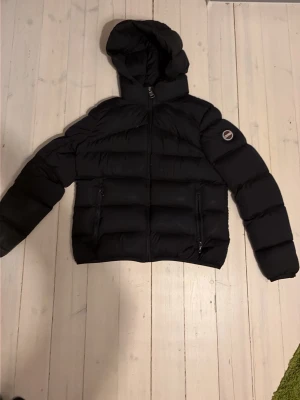 Svart pufferjacka med huva - Snygg svart pufferjacka med huva och dragkedja framtill. Jackan har två framfickor med dragkedja och ett runt märke på ena ärmen. Puffig design som ger en modern look och håller dig varm under kalla dagar.