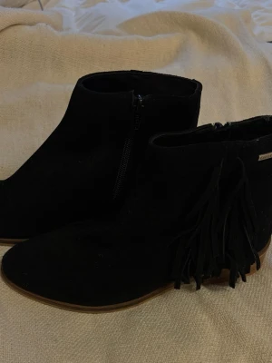Svarta stövletter med fransar - Snygga svarta stövletter från Firetrap i mocka med dekorativa fransar på sidan och låg klack. Skorna har dragkedja på insidan och en stilren, spetsig tå. Perfekta för dig som vill ha en trendig och cool look.