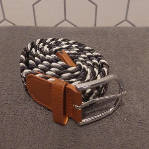 Flätat Bälte I Svart, Vit och Grå - Snyggt flätat bälte i svart, grå och vit med bruna läderdetaljer och ett stilrent silverfärgat spänne. Helt ny. Storlek 105 cm