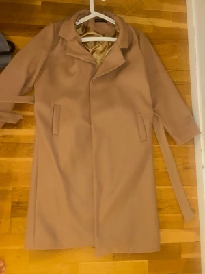 Klassisk beige kappa med skärp - Stilren beige kappa med knytband i midjan och två fickor framtill. Kappan har bred krage och är helfodrad med ljus insida. Perfekt för dig som gillar en enkel men elegant look. Materialet ser ut att vara ullblandning.